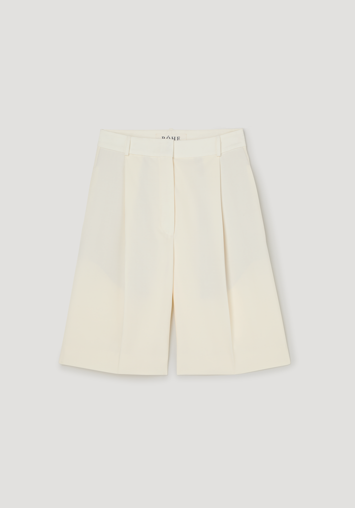Tailored single pleat shorts in off white, featuring knee-length, high-waisted design with front pleats, waistband with belt loops, and side pockets. Made from 100% virgin wool.