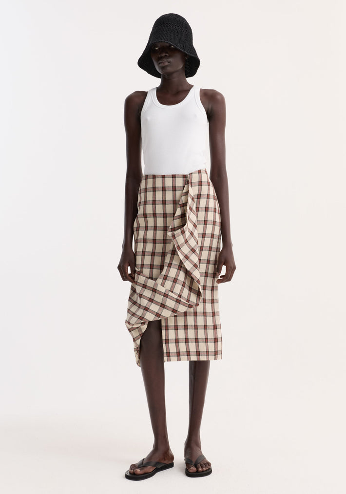 Model wearing a Draped check skirt | red table check, featuring knee-length, fitted silhouette with front ruffle drape, paired with a black crocheted hat and white tank top.