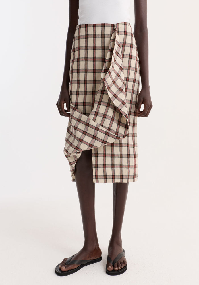 Model wearing Draped check skirt | red table check, featuring an asymmetrical front ruffle and knee-length fit, paired with a sleeveless top and sandals.