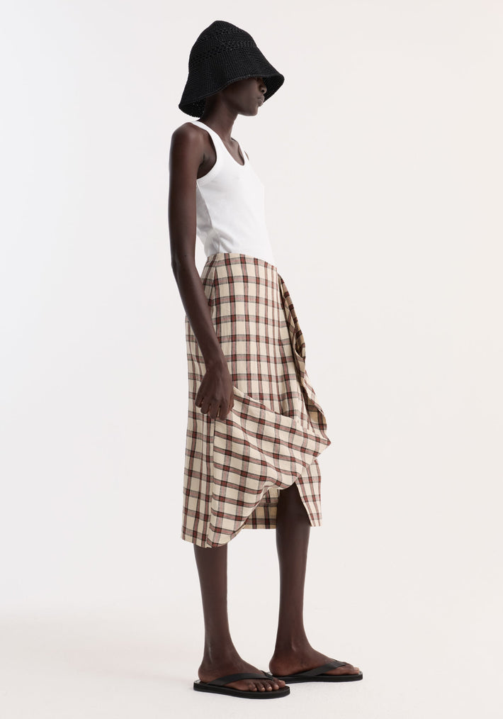 Model wearing Draped Check Skirt | Red Table Check in beige with red plaid, paired with a white tank top, black bucket hat, and black slide sandals.