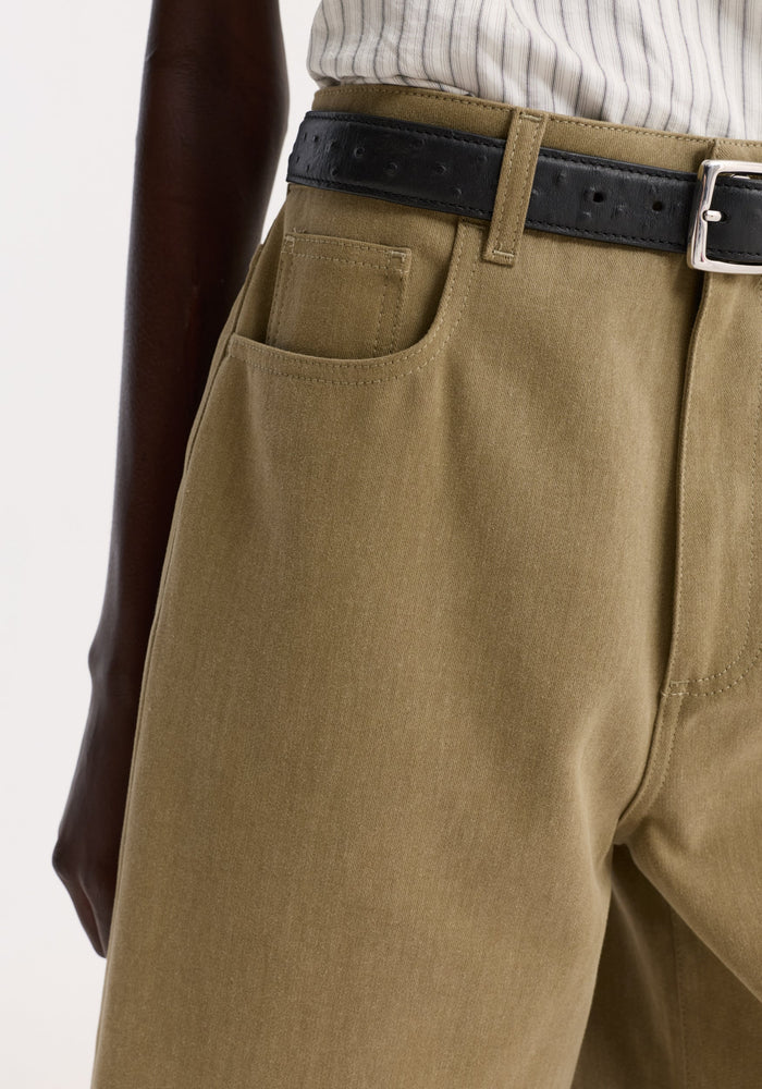 Close-up of baggy fit denim shorts in sage, showcasing Bermuda length and subtle tone variations, worn with a black leather belt and tucked-in striped shirt.