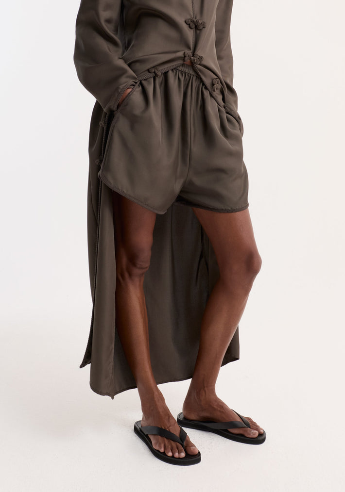 Model wearing Taped silk shorts | earth, featuring fluid silk, elasticated waistband, and embroidered hem, styled with a high-low top and black flip-flops.
