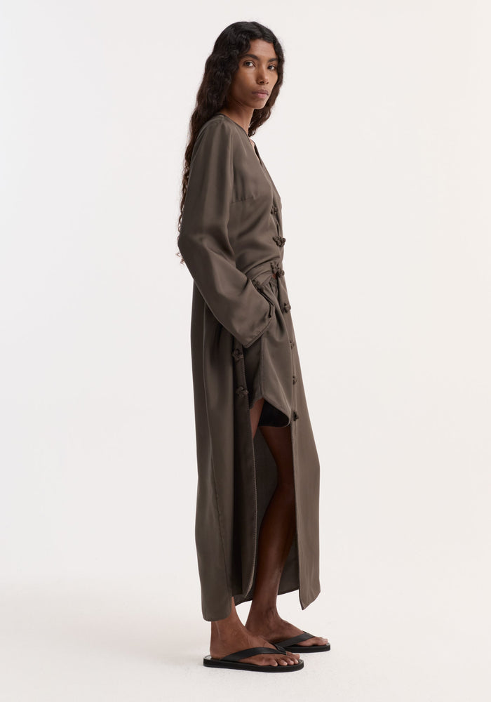 Model in profile wearing olive-brown duster, revealing Taped silk shorts | earth with elastic waistband and pleated, embroidered hem. Complemented by black flat sandals.
