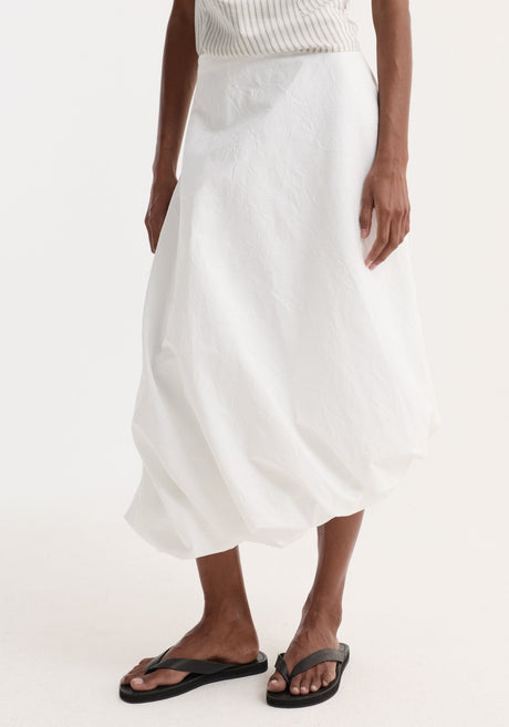 Model wearing Twisted crushed cotton skirt | optic white, featuring high-waisted, voluminous balloon hem, paired with a pinstriped top, black tote, and sandals.