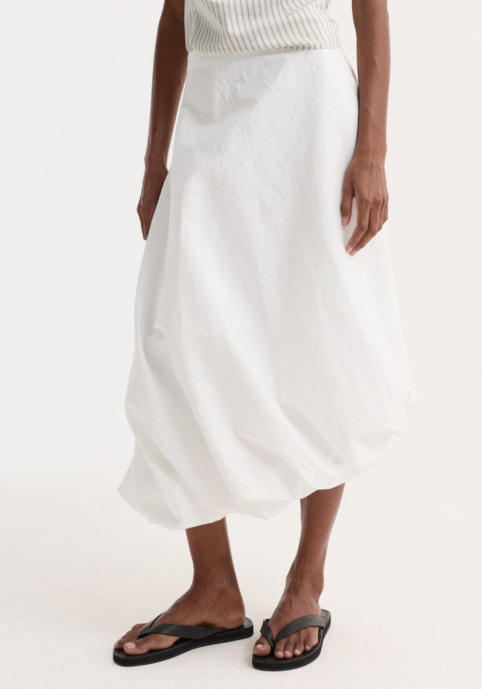 Model wearing Twisted crushed cotton skirt in optic white, featuring a voluminous bubble design and twist detail at hem, paired with black flip-flop sandals.