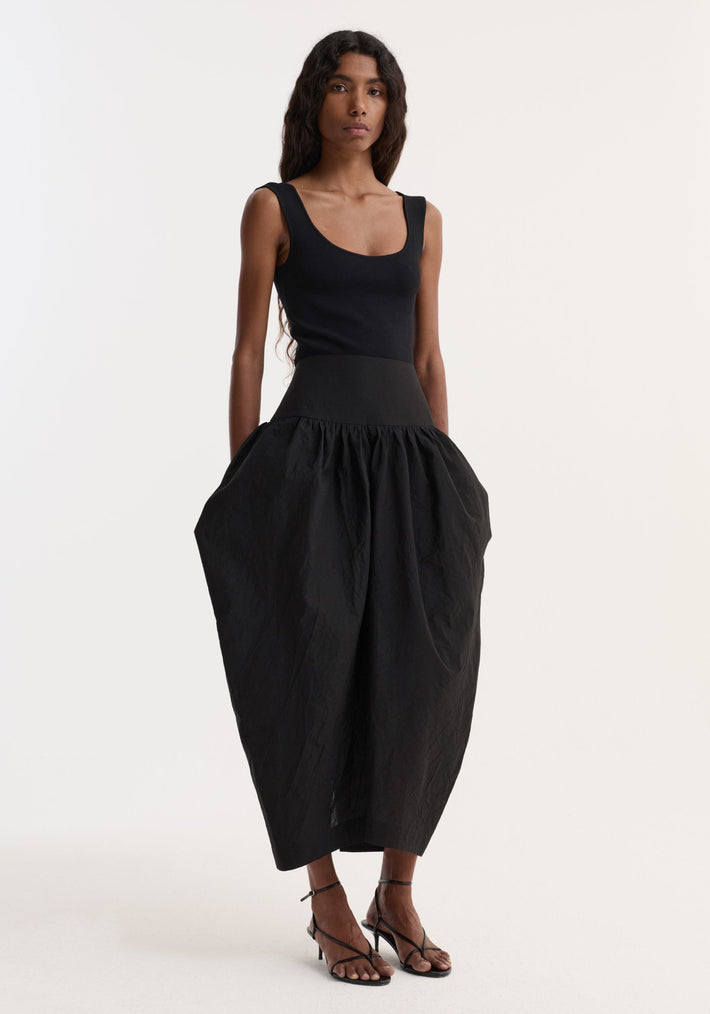 Model wearing the Sculptural volume skirt | black, featuring a mid-waist, fitted waistband, and architectural pockets, paired with a sleeveless top and strappy heeled sandals.