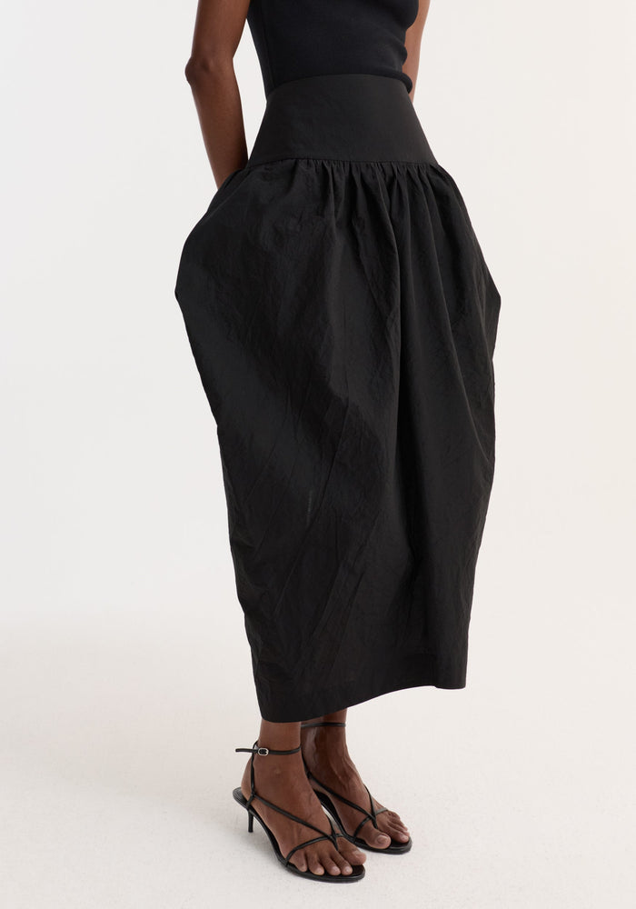 Model wearing Sculptural volume skirt | black, featuring a high-waisted, voluminous silhouette with architectural pockets, paired with strappy black sandals in a studio setting.