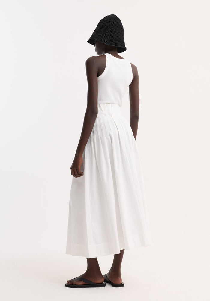 Rear view of model in a Pleated poplin maxi skirt, optic white, showcasing its high-waist pleated design with wide hem, paired with minimalist accessories.