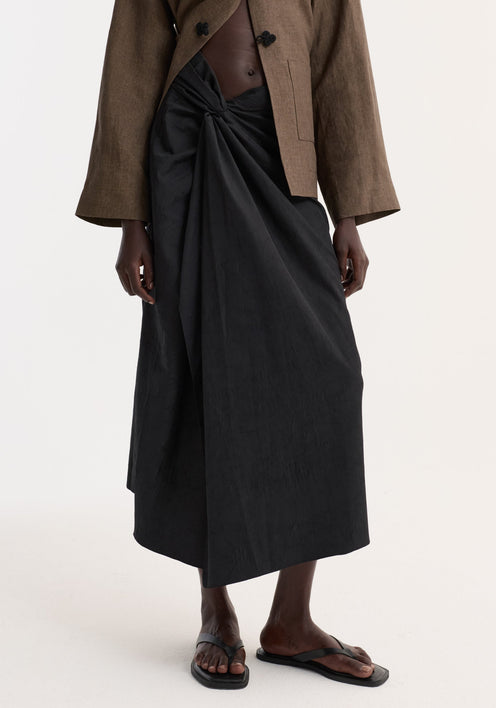 Model wearing Sarong textured skirt | black, featuring a front knot and gathered waistband. Paired with a cropped jacket, showcasing the skirt's elegant drape and style.