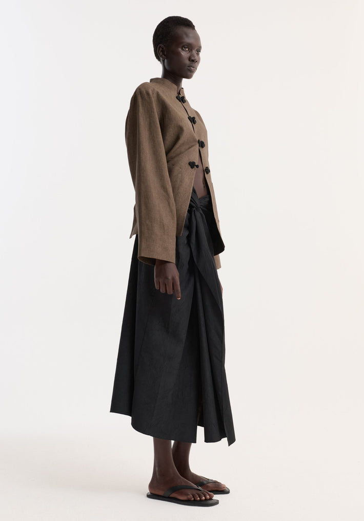 Model in profile wearing Sarong textured skirt | black with knot detail, draped design, and concealed zip, paired with a cropped jacket and flat sandals.