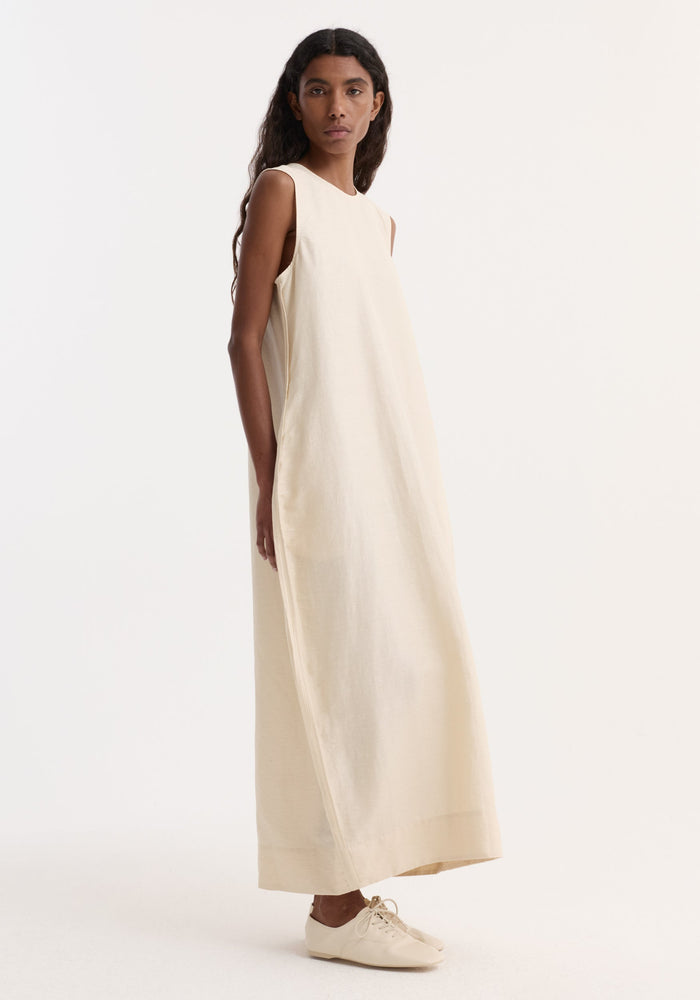 Woman in Hourglass linen silk dress | butter, showcasing a sculptural, sleeveless floor-length design with a softly crushed texture, paired with matching lace-up flats.