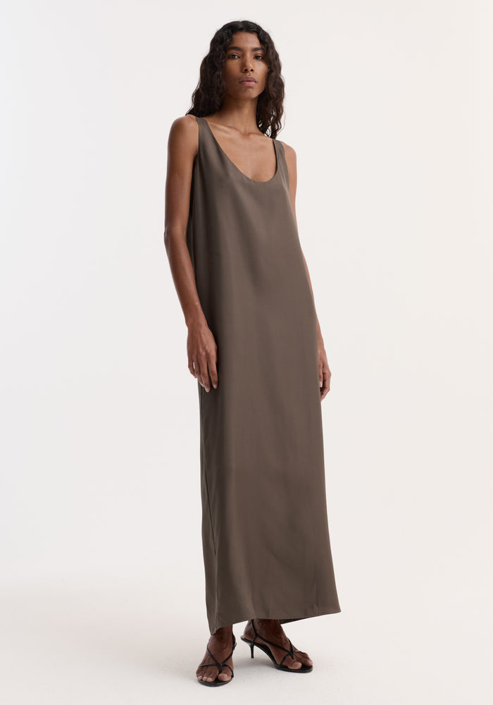 Model in an olive-taupe Silk scoop-neck dress | earth, featuring a sleeveless, floor-length design in 100% silk, paired with black strappy sandals.