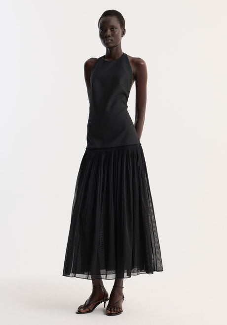Model wearing Pleated muslin halter dress | black with a structured bodice, low back, and sheer pleated skirt, paired with black strappy sandals.