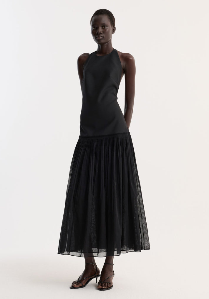 Model wearing Pleated muslin halter dress | black with a structured bodice, low back, and sheer pleated skirt, paired with black strappy sandals.
