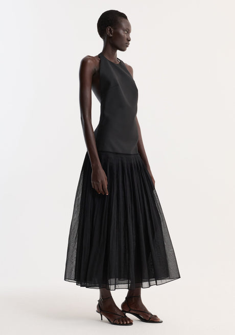 Model wearing Pleated muslin halter dress | black with a structured bodice, low back, and sheer pleated skirt, paired with black strappy sandals.