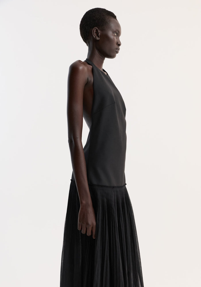 Pleated muslin halter dress in black, featuring a structured bodice and low back, worn by a model standing in profile with a flowing pleated skirt.