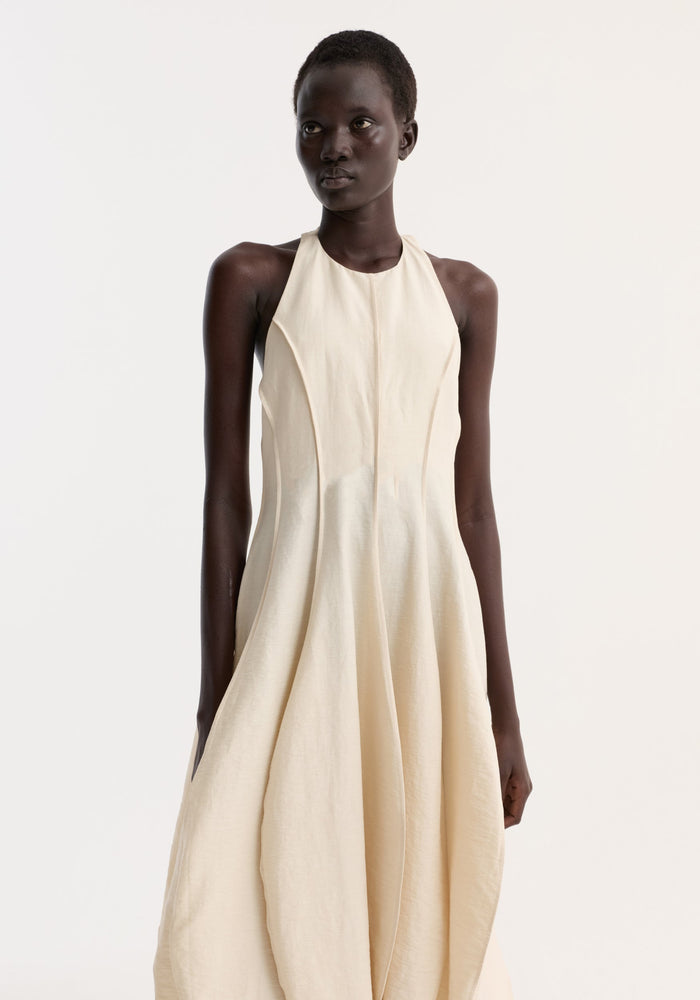 Model in a halter-neck Open back tulip dress | butter, featuring a fitted bodice, wide pleats, balloon-shaped skirt, high neckline, and low back.