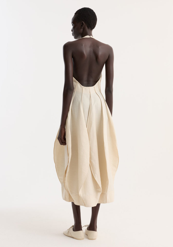 Model in Open back tulip dress | butter, showcasing a cream backless halter midi dress with draped petal-like panels and pleated balloon-shaped skirt, paired with neutral lace-up shoes.