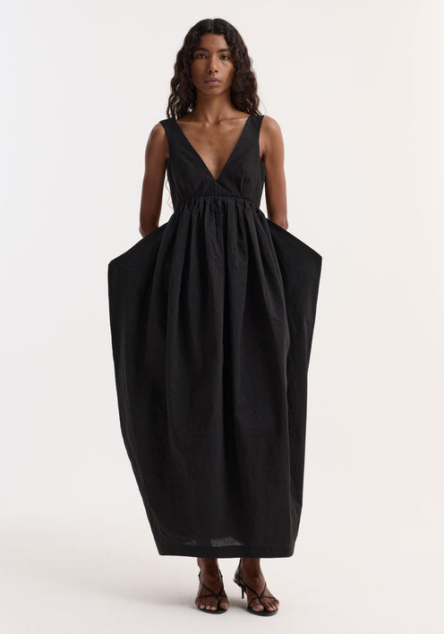 Woman in Sculptural textured cotton dress | black, featuring a deep-V neckline, low back, voluminous skirt, and discreet pockets, paired with black strappy heeled sandals.