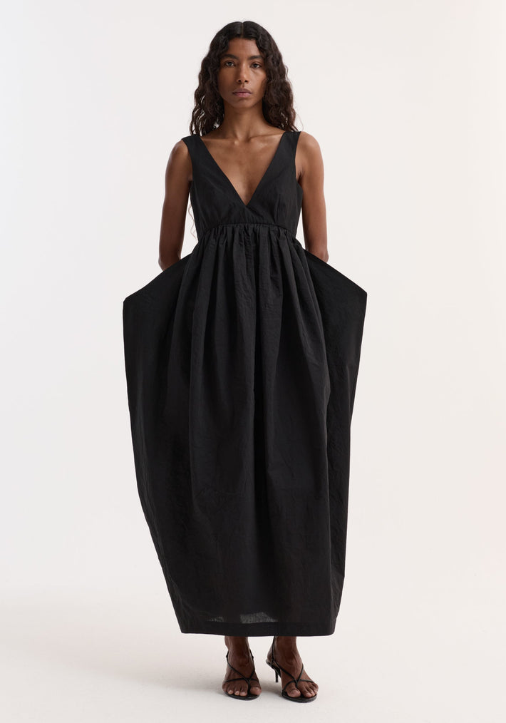 Woman in Sculptural textured cotton dress | black, featuring a deep-V neckline, low back, voluminous skirt, and discreet pockets, paired with black strappy heeled sandals.
