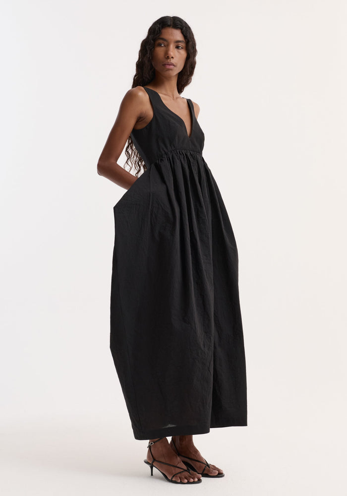 Model in profile wearing the Sculptural textured cotton dress | black, featuring a sleeveless V-neck, gathered waist, trapezium-shaped skirt, and side pockets.