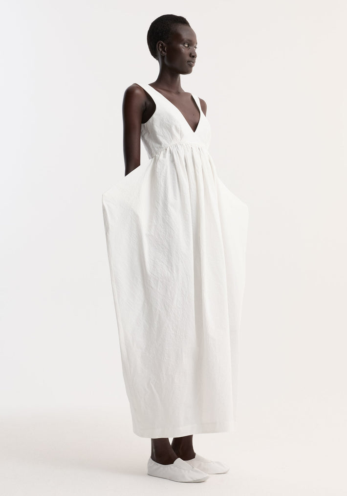 Model in profile wears Sculptural textured cotton dress, off white, with sleeveless V-neck, full billowy skirt, and side-seam pockets, showcasing a trapezium silhouette.