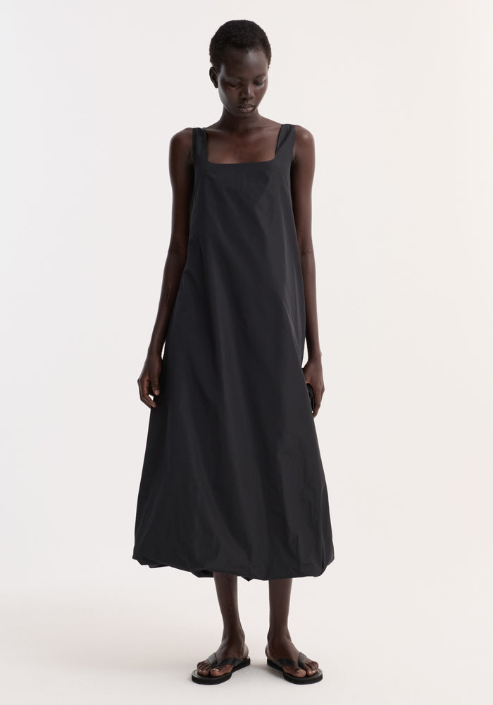 Tall woman in a Technical volume dress | black, featuring wide straps, scooped square neckline, and balloon hemline, paired with black slide sandals.