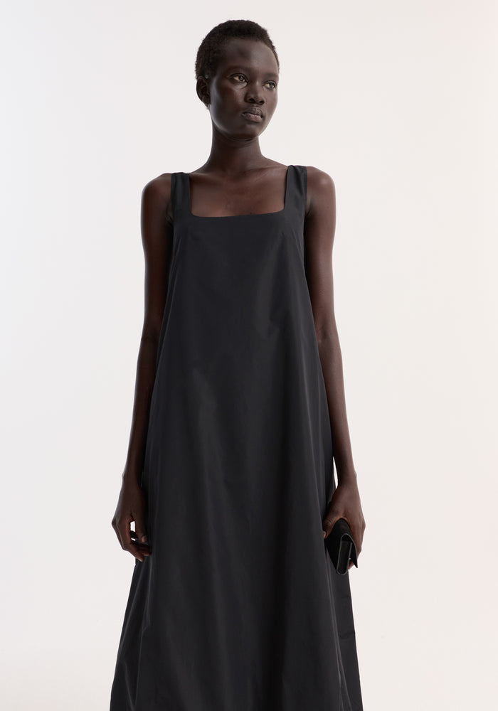 Model in Technical Volume Dress, showcasing a sleeveless design with wide straps, a square neckline, and a balloon hemline, holding a small clutch.