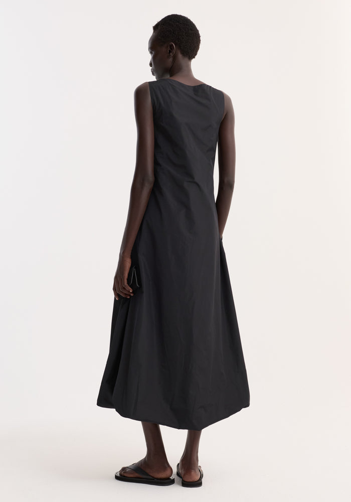 Dark-skinned woman models the Technical volume dress in black, showcasing its sleeveless design, square neckline, and balloon hemline, paired with black sandals.