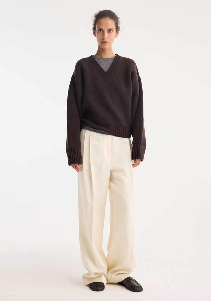 Model wearing Sculptural compact V-neck in dark brown, featuring a chunky knit design, relaxed fit, and Italian wool craftsmanship, paired with wide-leg cream trousers.