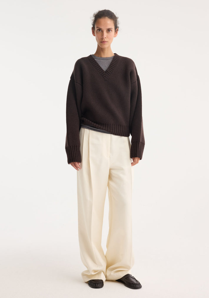Model wearing Sculptural compact V-neck in dark brown, featuring a chunky knit design, relaxed fit, and Italian wool craftsmanship, paired with wide-leg cream trousers.