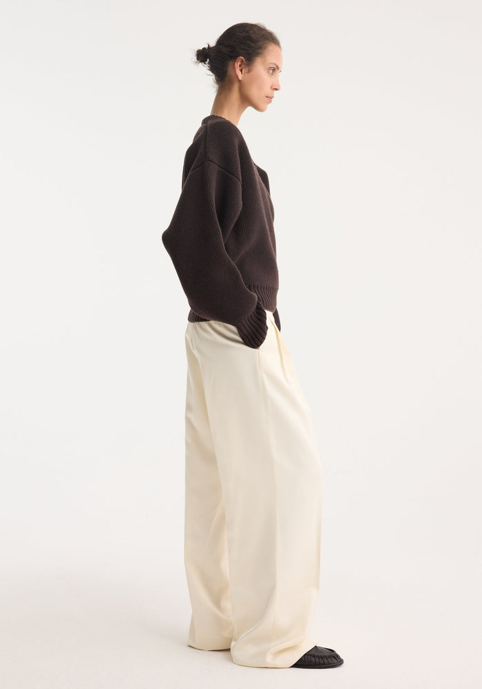 Model wearing the Sculptural compact v-neck in dark brown, paired with high-waisted wide-leg trousers and flat slip-on shoes, showcasing a relaxed minimalist style.