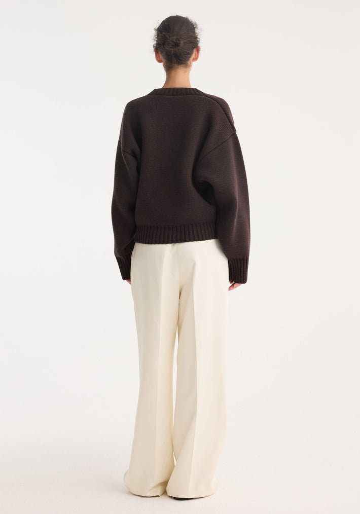 Person wearing Sculptural compact V-neck dark brown sweater with dropped shoulder and ribbed details, paired with high-waisted, wide-leg cream trousers, hair in a bun.