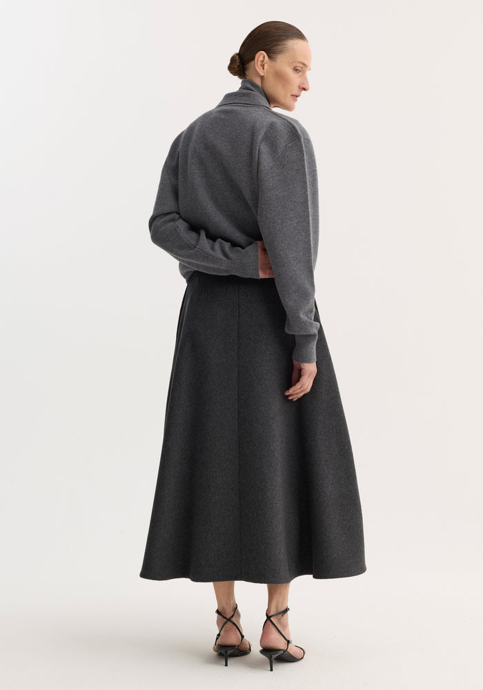 Woman wearing an anthracite melange A-line skirt, showcasing its mid-length, voluminous silhouette, paired with a gray pullover and black sandals, viewed from behind.