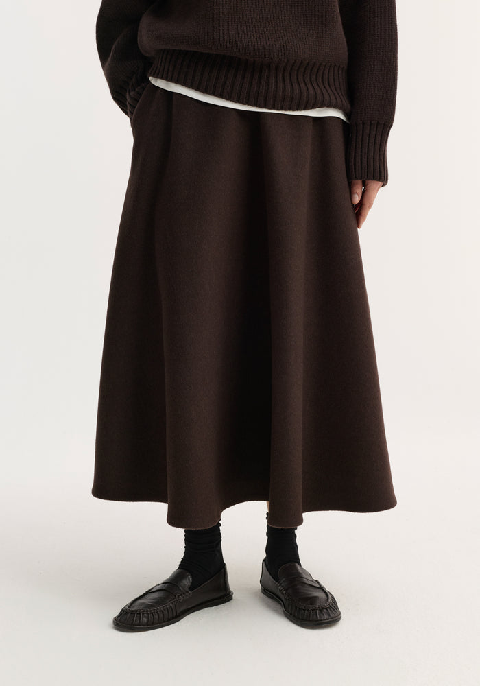 A-line skirt | dark brown: Model wears a voluminous A-line, ankle-length skirt with a textured surface, paired with a matching knit sweater, black socks, and loafers.
