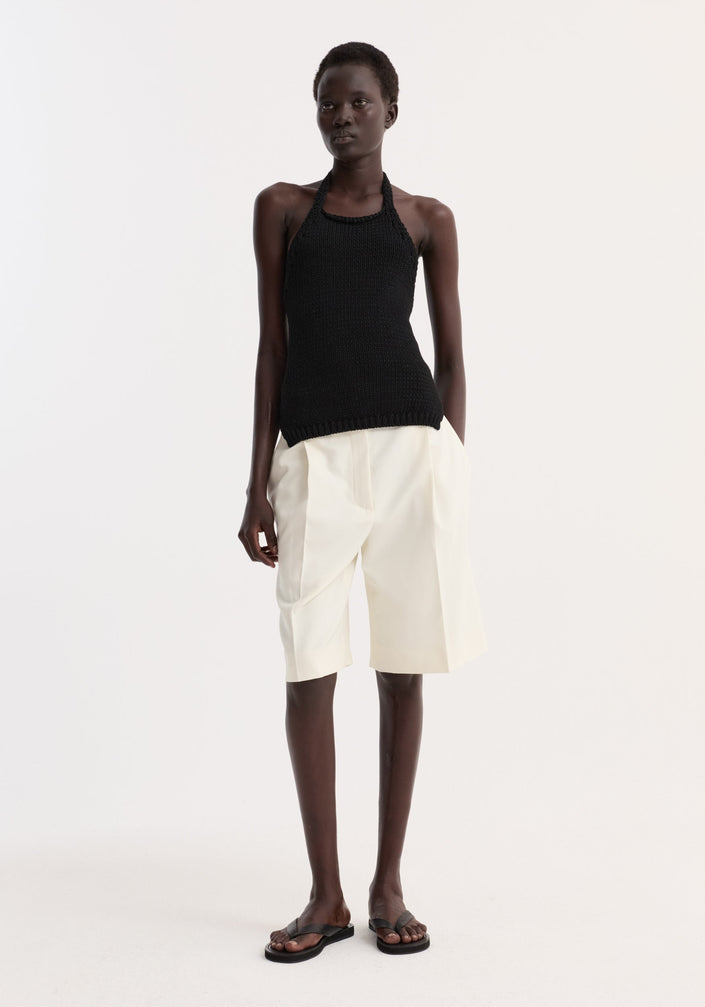 Model wears Chunky knit halter top | black, a sleeveless, hand-knit cotton design with ribbed hem and button closure, paired with pleated Bermuda shorts and flat sandals.