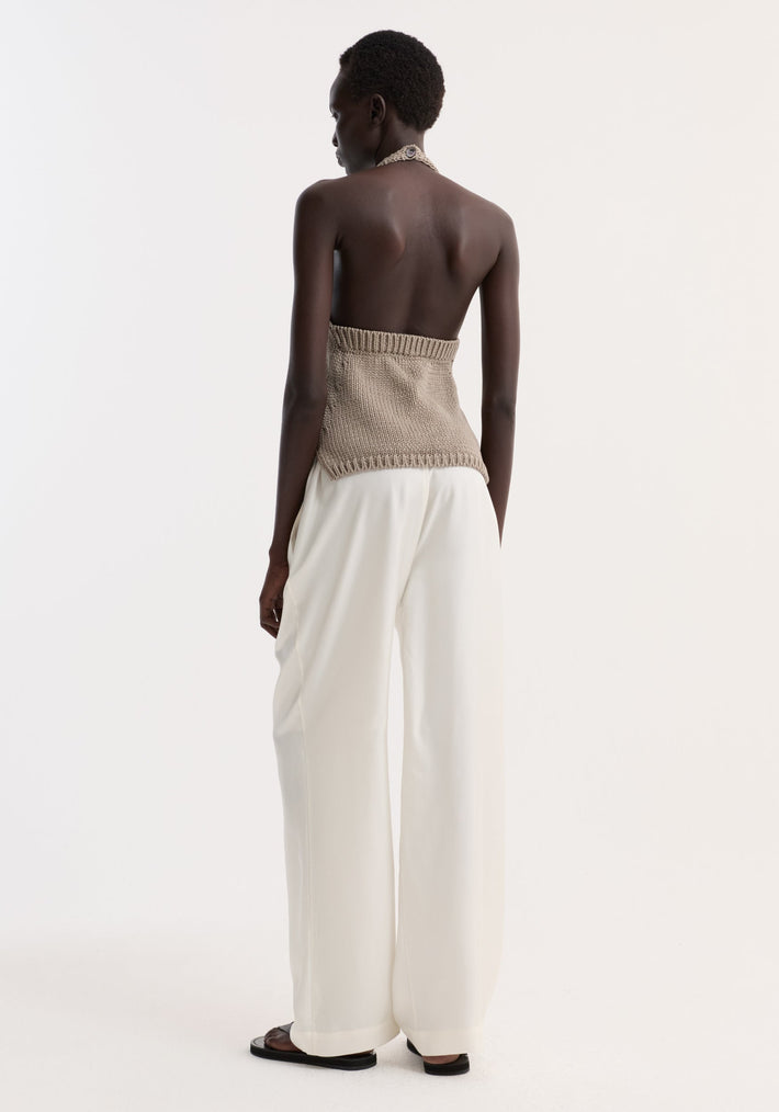 A tall woman models the Chunky knit halter top | stone with side slits, button neck closure, paired with high-waisted white trousers.