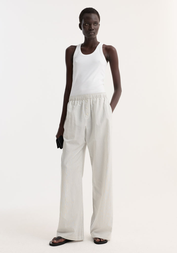 Silk stripe pyjama trousers with fine vertical stripes, wide-leg fit, and elasticated waistband, worn by a woman with black sandals and a clutch.