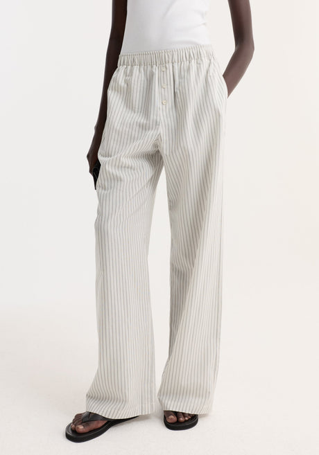 Person wearing Silk stripe pyjama trousers with fine vertical stripes, high-waisted, wide-leg design, and elasticated waistband, paired with a white tank top and black flip-flops.