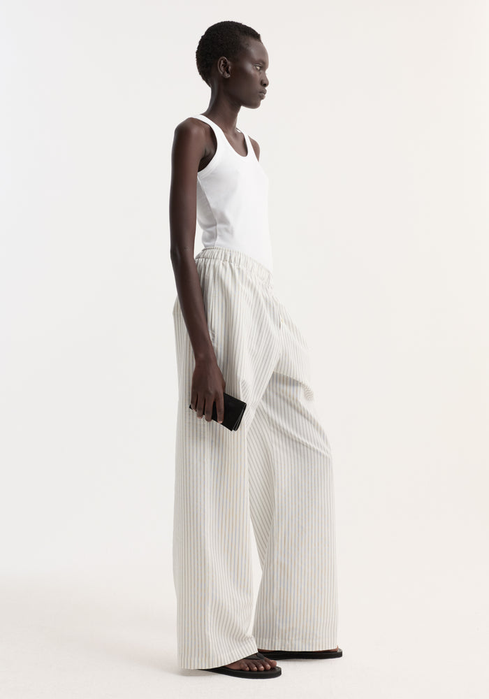 Model in profile wearing Silk stripe pyjama trousers with wide-leg design, elasticated waistband, and faux fly, paired with a white ribbed tank top and black sandals.