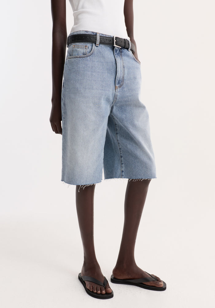 Baggy fit denim shorts in classic blue with a raw-edge hem, worn with a black belt and flip-flops, showcasing a relaxed, casual style.