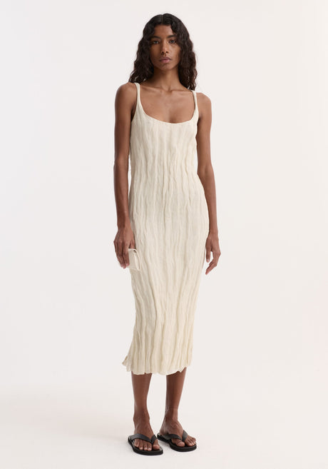 Model in a textured square neck dress | cream, with metal yarn crinkles, holding a matching purse, wearing black sandals, posed against a white backdrop.