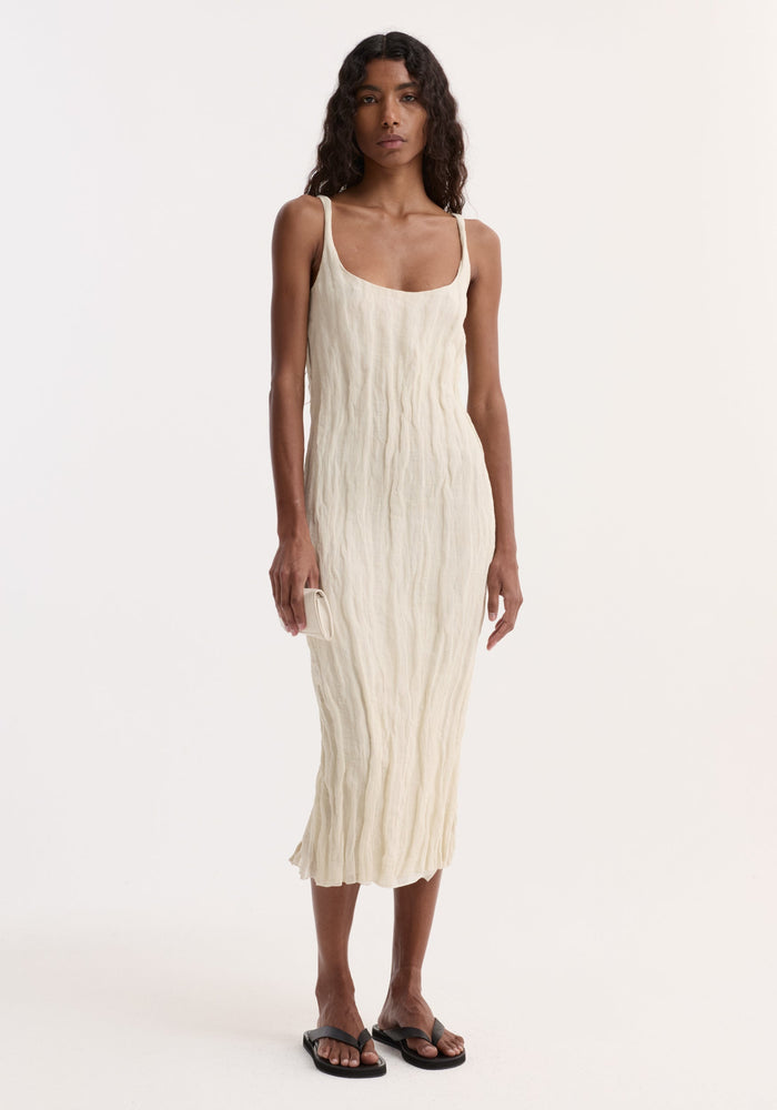Model in a textured square neck dress | cream, with metal yarn crinkles, holding a matching purse, wearing black sandals, posed against a white backdrop.