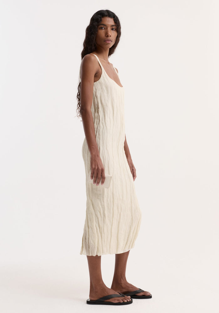Woman in a Textured square neck dress | cream, sleeveless with a scoop neck, holding a small item, showcasing the dress's crinkled texture and straight silhouette.