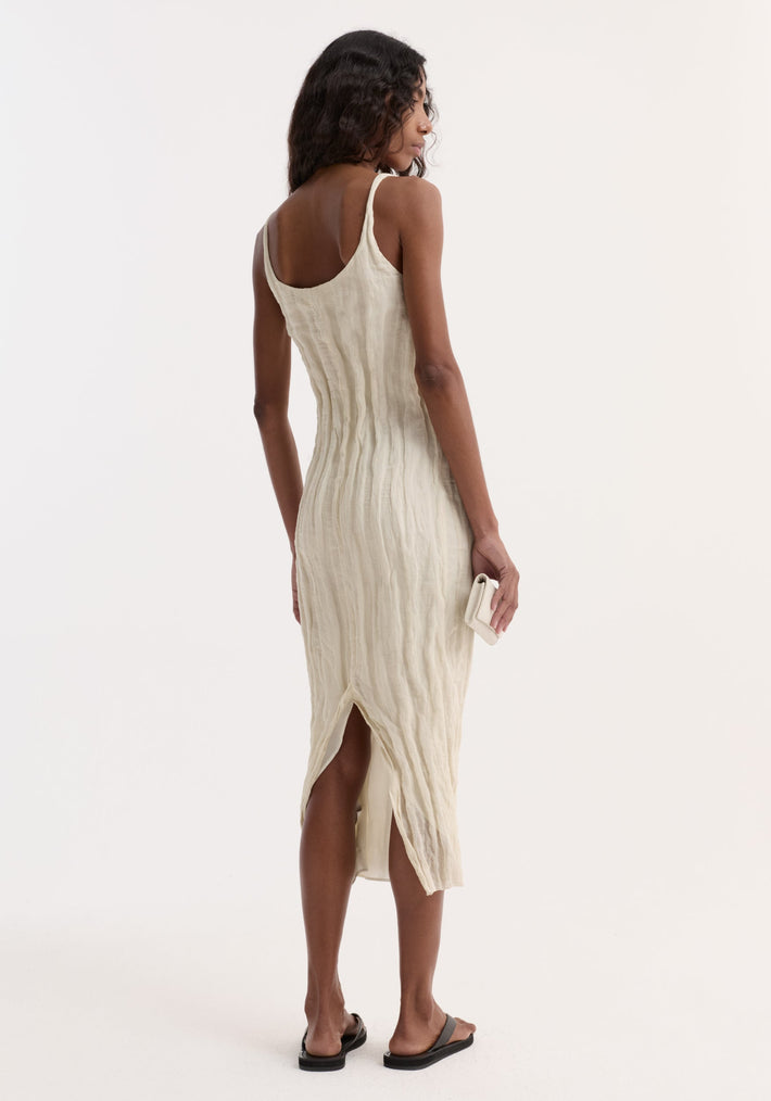 Textured square neck dress in cream, three-quarter rear view. Features sleeveless design, crinkle texture, small back slit. Paired with clutch and black slide sandals.