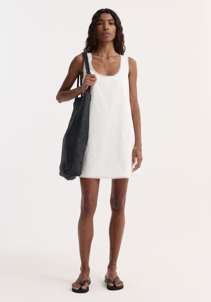Model in A-line poplin mini dress, optic white, with scalloped trim, carrying black tote. Wearing black sandals. Dress has embroidery at neckline and hem.