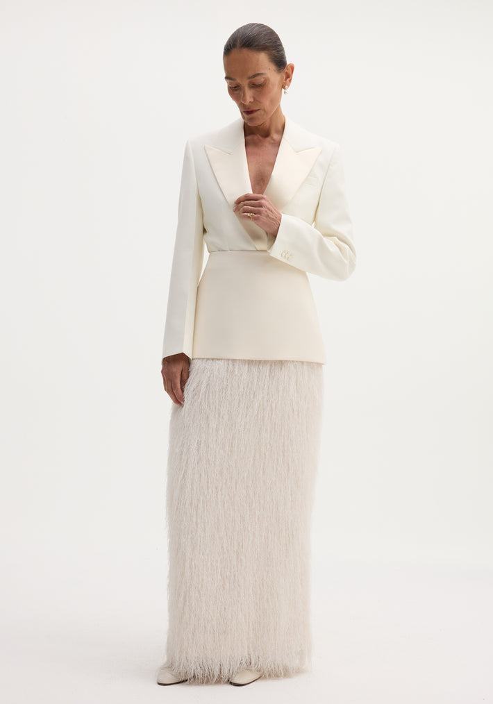 Model in an ivory Fringe skirt featuring a fitted wool upper and full-length fringe, paired with a tailored blazer, pearl earrings, and slicked-back hair.