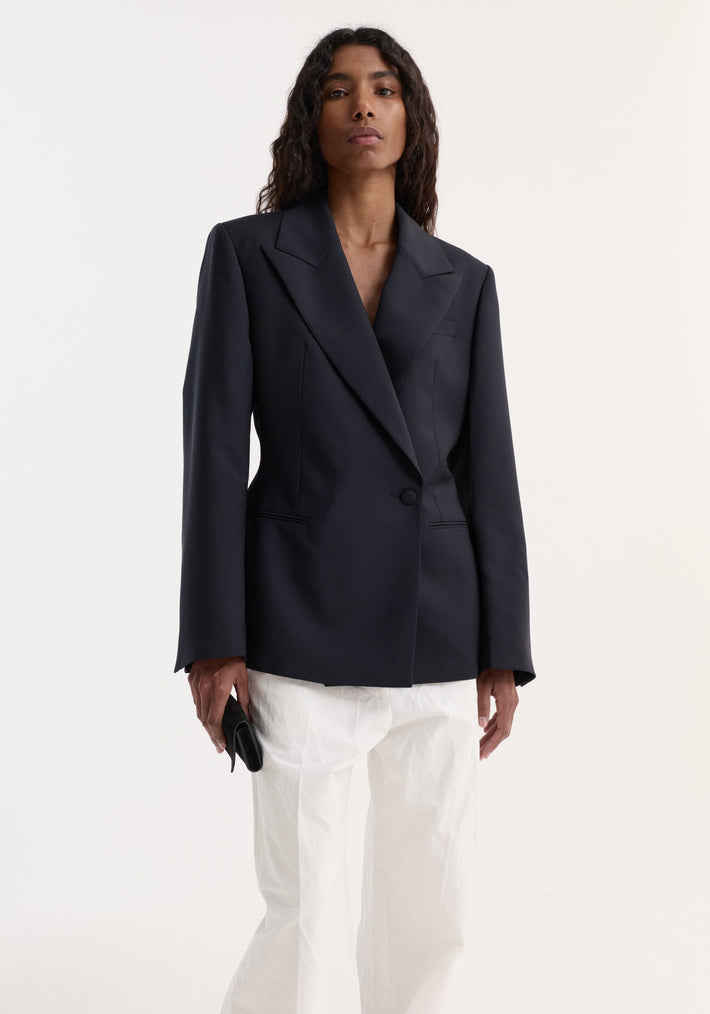 Model in Hourglass shaped blazer with sharp notch lapel, cinched waist, single button closure, paired with loose-cut trousers, holding a black clutch.