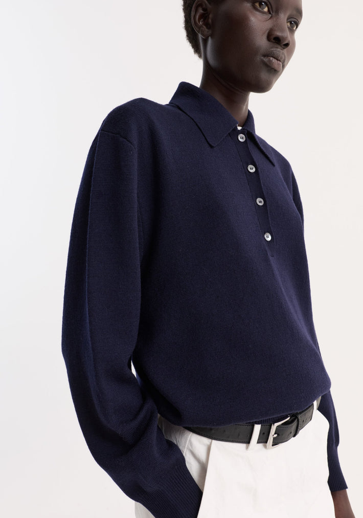 Model wearing a navy Merino wool polo sweater with a structured collar and four-button placket, tucked into light trousers with a black leather belt.