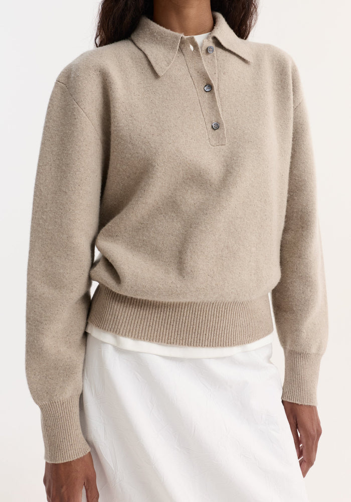 Person wearing Wool cashmere polo | stone melange, featuring a light-beige knit with a polo collar, three-button placket, ribbed cuffs, and hem. White skirt visible below.